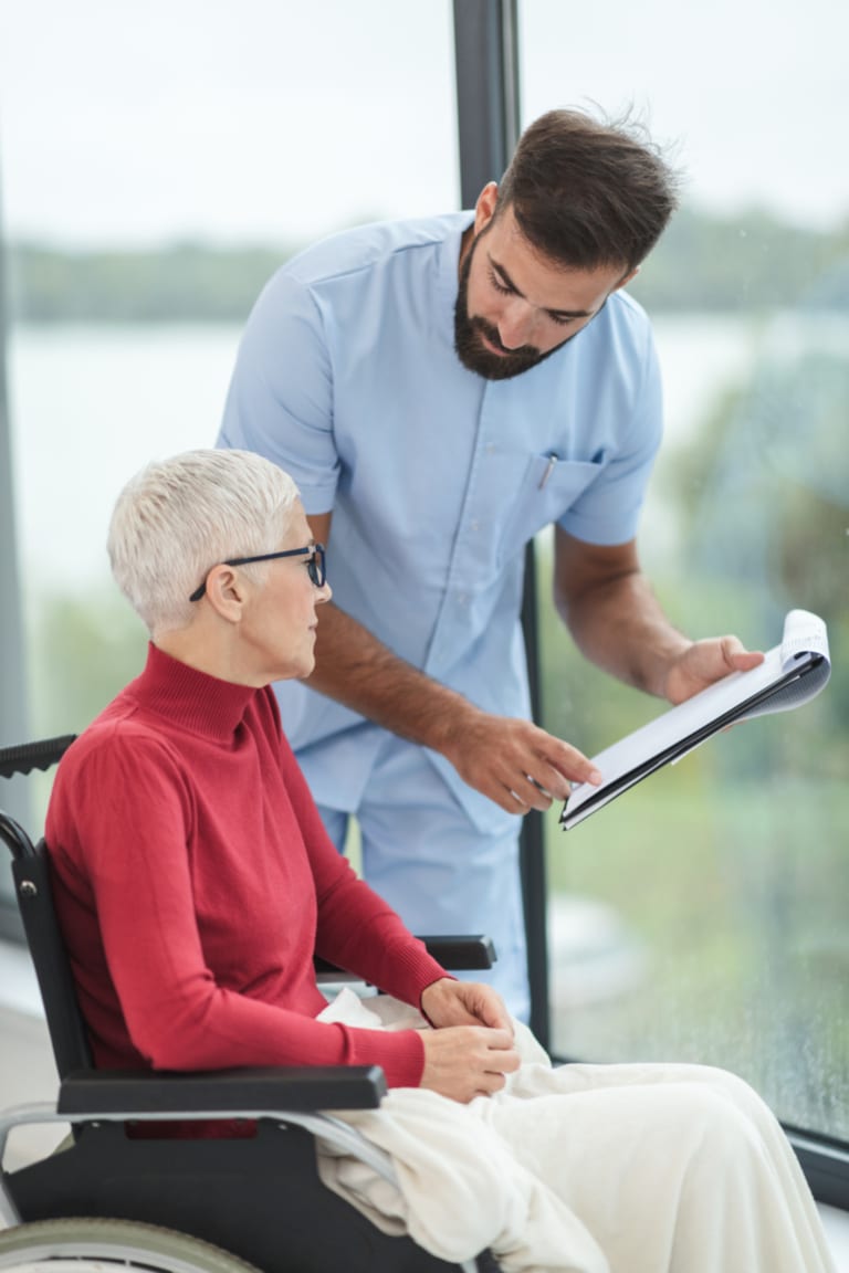 A caregiver reviewing a care plan on a clipboard with an older woman by a window.