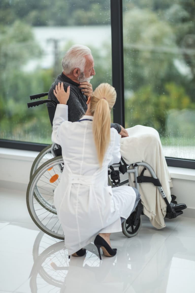 A clinician kneeling beside an older man in a wheelchair near a garden window.