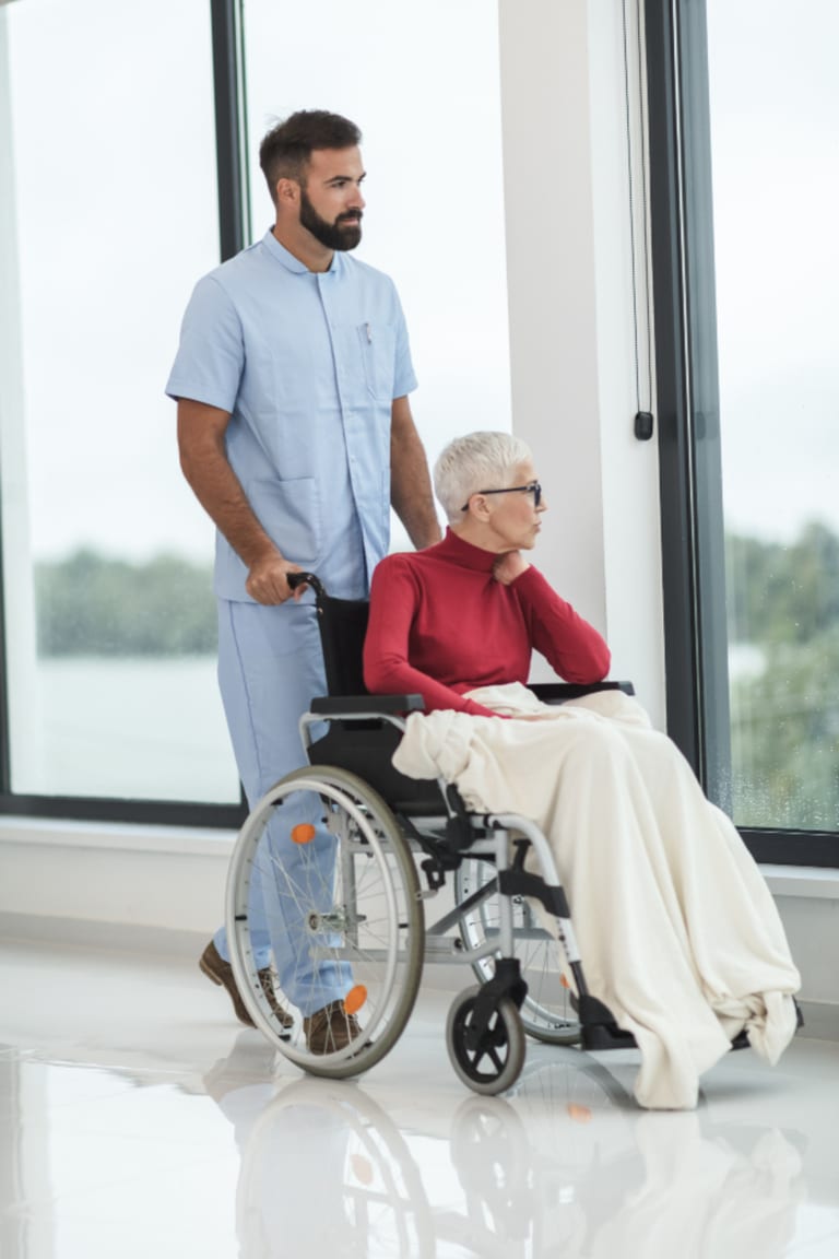 A caregiver standing with an older woman in a wheelchair looking out a large window.
