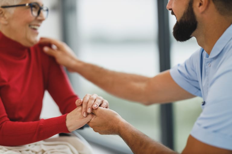 A caregiver gently holding hands with a smiling older woman in a bright home setting.