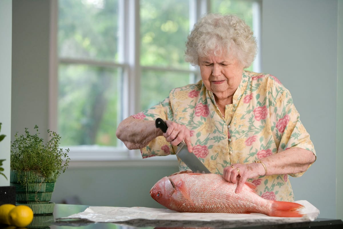 An older woman preparing fresh food at a sunlit home kitchen counter.